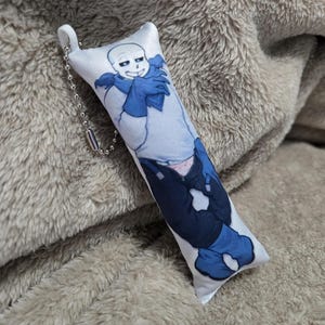 May include: A white rectangular plush keychain featuring a cartoon character with a skull-like face, blue scarf, and blue clothing. The keychain has a silver chain and a white loop for attachment.