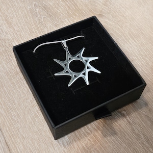 May include: A silver-toned metal pendant shaped like a stylized sun with eight sharp, pointed rays and a circular center cutout. The pendant is attached to a thin silver-toned chain and is presented inside a square black jewelry box with a velvet lining.