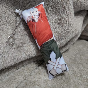 May include: A rectangular plush keychain featuring a cartoon character in a red hoodie and green shorts. The keychain has a silver chain and is resting on a soft, gray surface.