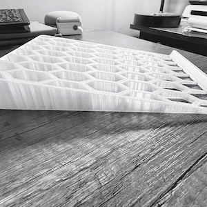 May include: A white, wedge-shaped stand featuring a repeating hexagonal grid pattern across its top surface. The object has a sloped profile, rising from a thin front edge to a thicker back edge, constructed with a visible 3D printed texture.