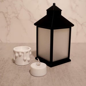 May include: A black lantern with a white, frosted glass panel, a white candle, and a white candle holder with a dripping wax design. The lantern has a square base and a peaked roof.