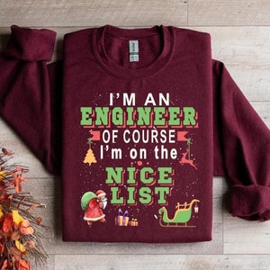 May include: A maroon sweatshirt featuring the text "I'M AN ENGINEER OF COURSE I'm on the NICE LIST" in green and white. The design includes festive Christmas imagery such as Santa and a sleigh.