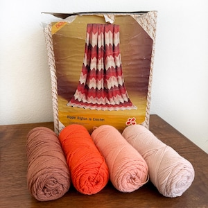 May include: A vintage crochet kit with a box featuring a red, white, and brown chevron afghan design. Four yarn skeins in shades of brown, orange, and pink are in front of the box. The box reads "Ripple Afghan to Crochet."