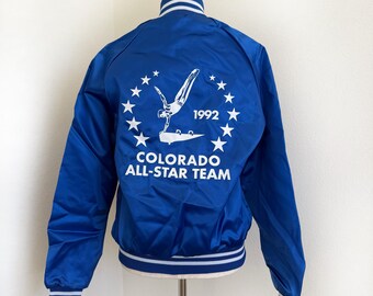 May include: A royal blue satin jacket with white trim on the collar, cuffs, and hem. The back features a white graphic of a gymnast and the text "COLORADO ALL-STAR TEAM 1992" surrounded by stars. The jacket is displayed on a mannequin.