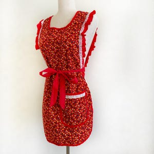 May include: A vintage-style red apron with a floral pattern, white lace trim, and a red bow. The apron features a pocket and ruffled edges. The apron is displayed on a mannequin against a white background.