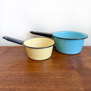 May include: Two vintage enamelware saucepans. One is light yellow with a black handle and rim, the other is light blue with a black handle and rim. Both pans are on a wooden surface.