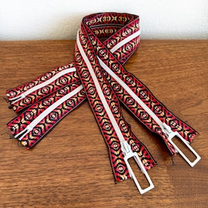 May include: Decorative zipper with a geometric pattern in red, black, and yellow, with a white stripe down the center. The zipper has a silver-colored pull and is displayed on a wooden surface. This item is suitable for sewing and craft projects.