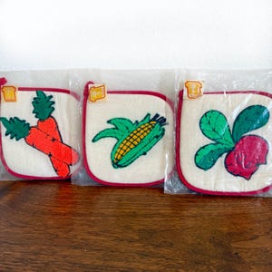 May include: Three square, off-white pot holders with embroidered vegetable designs: carrots, corn, and a radish. Each has a red border and a small tag. The pot holders are packaged in clear plastic.