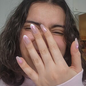 May include: Close-up of a hand with pink and white oval-shaped nails with a white tip and pink heart design. The hand is partially covering the face of a person with long dark hair.