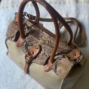 Może przedstawiać: Beżowa skórzana torebka z wężowym wzorem na przedniej klapie. Torebka ma dwa górne uchwyty i długi pasek na ramię.