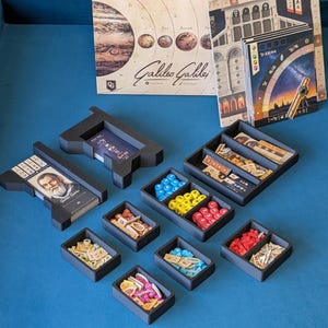 May include: A board game titled "Galileo Galilei" with various game components, including cards, tokens, and black storage trays. The game box features images of planets and historical figures. The game is set on a blue surface.