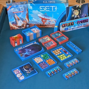 May include: A board game titled "SETI: Search for Extraterrestrial Intelligence" with a space theme. The game includes boxes, cards, and various game pieces in blue, red, and other colors. The game is set on a blue surface.