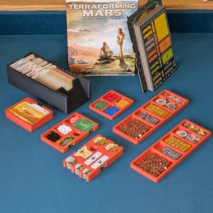 May include: A board game titled "Terraforming Mars: Ares Expedition" with various components. Includes a black card holder, red trays with game pieces, and a rulebook. The game pieces are in various colors and shapes.