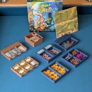 May include: Everdell Duo board game set featuring a colourful game box, a square game board, and several brown and blue plastic organisers filled with various game components, including small cards, circular tokens, and miniature animal figures.
