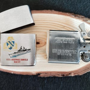 Puede incluir: Un encendedor Zippo plateado con la tapa abierta, revelando el interior. El encendedor est&aacute; decorado con una ilustraci&oacute;n de un barco y el texto "USS JOSEPHUS DANIELS DLG-27". La otra parte del encendedor tiene el texto "USE ZIPPO FLINTS AND FLUID".