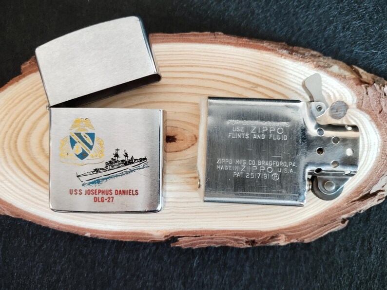 Puede incluir: Un encendedor Zippo plateado con la tapa abierta, revelando el interior. El encendedor est&aacute; decorado con una ilustraci&oacute;n de un barco y el texto "USS JOSEPHUS DANIELS DLG-27". La otra parte del encendedor tiene el texto "USE ZIPPO FLINTS AND FLUID".