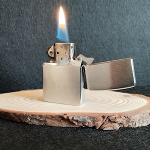 Puede incluir: Un encendedor de metal plateado con la tapa abierta y una llama encendida. La llama es amarilla y azul. El encendedor est&aacute; sobre una rodaja de madera redonda con un fondo oscuro.