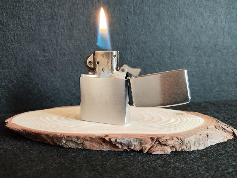 Puede incluir: Un encendedor de metal plateado con la tapa abierta y una llama encendida. La llama es amarilla y azul. El encendedor est&aacute; sobre una rodaja de madera redonda con un fondo oscuro.