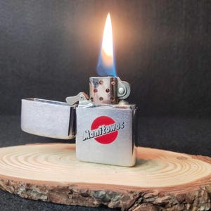 Peut inclure: Un briquet argent&eacute; avec une flamme allum&eacute;e, pos&eacute; sur une tranche de bois. Le briquet porte l'inscription "Manitowoc" et un cercle rouge. La flamme est jaune et bleue.