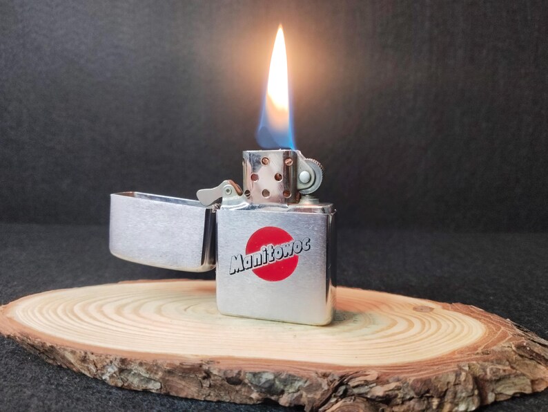 Peut inclure: Un briquet argent&eacute; avec une flamme allum&eacute;e, pos&eacute; sur une tranche de bois. Le briquet porte l'inscription "Manitowoc" et un cercle rouge. La flamme est jaune et bleue.