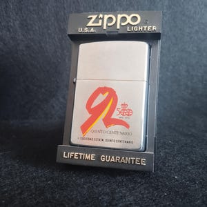 Puede incluir: Un encendedor Zippo plateado en su embalaje negro original. El encendedor presenta un diseño rojo y amarillo con el texto "QUINTO CENTENARIO" y "500 AÑOS 1492-1992". El embalaje dice "ZIPPO U.S.A. LIGHTER" y "LIFETIME GUARANTEE".