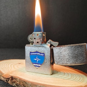 Może przedstawiać: Srebrna metalowa zapalniczka z niebiesko-białym emblematem z napisem "STANAVFORLANT" i symbolem NATO. Zapalniczka jest zapalona, z pomarańczowo-niebieskim płomieniem. Zapalniczka spoczywa na drewnianym plastrze.