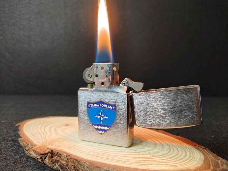 Może przedstawiać: Srebrna metalowa zapalniczka z niebiesko-białym emblematem z napisem "STANAVFORLANT" i symbolem NATO. Zapalniczka jest zapalona, z pomarańczowo-niebieskim płomieniem. Zapalniczka spoczywa na drewnianym plastrze.