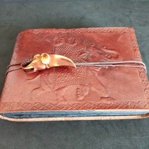 Puede incluir: Un diario encuadernado en cuero marrón con un diseño de elefante en relieve. El diario tiene un cierre decorativo en forma de colmillo de elefante.