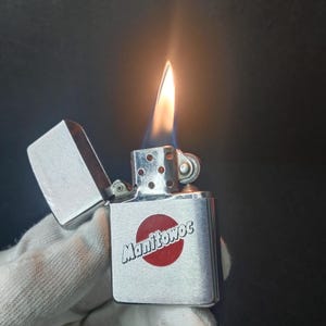 Peut inclure: Un briquet en m&eacute;tal argent&eacute; avec une flamme allum&eacute;e. Le briquet porte le mot "Manitowoc" en blanc sur un cercle rouge. Le briquet est tenu par une main gant&eacute;e.