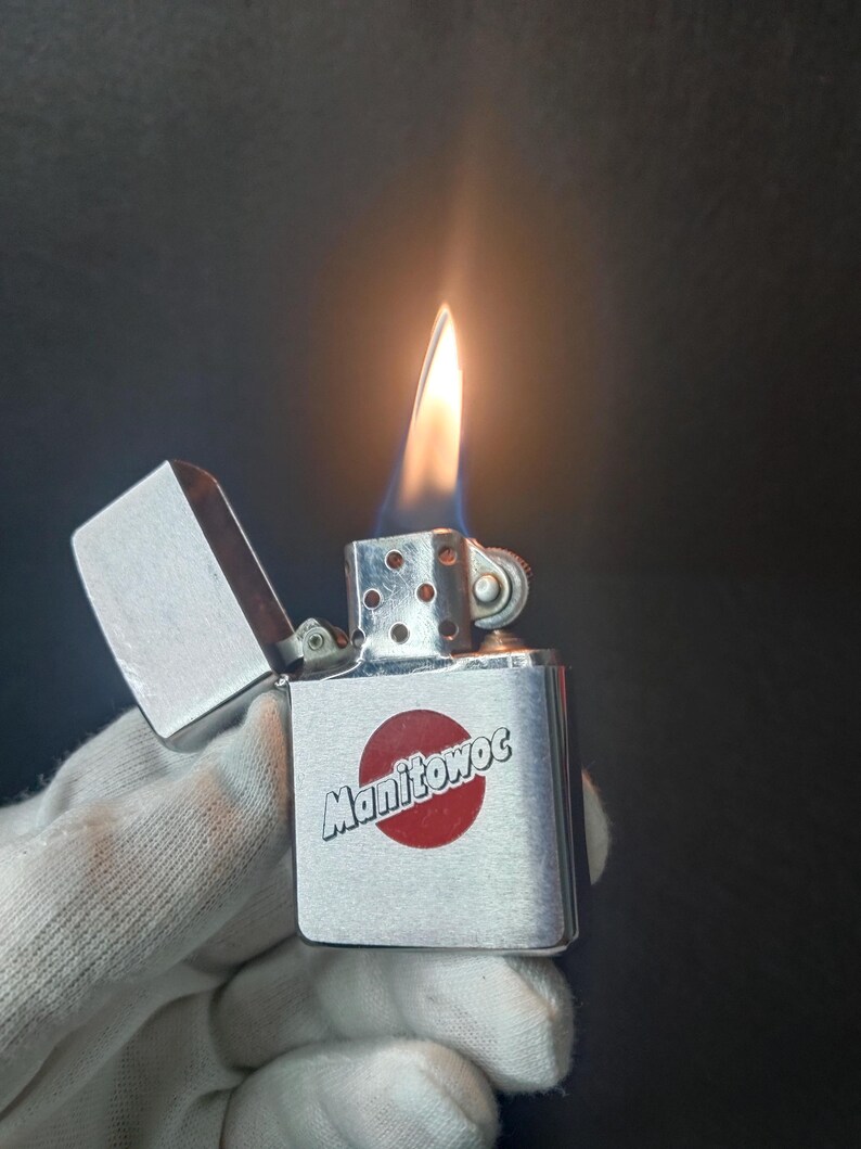 Peut inclure: Un briquet en m&eacute;tal argent&eacute; avec une flamme allum&eacute;e. Le briquet porte le mot "Manitowoc" en blanc sur un cercle rouge. Le briquet est tenu par une main gant&eacute;e.