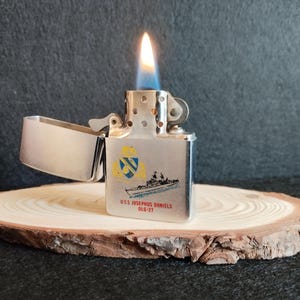 Puede incluir: Un encendedor de metal plateado con una llama, sobre una rodaja de madera. El encendedor presenta un dise&ntilde;o de barco y el texto "USS JOSEPHUS DANIELS DLG-27" en negro y azul.