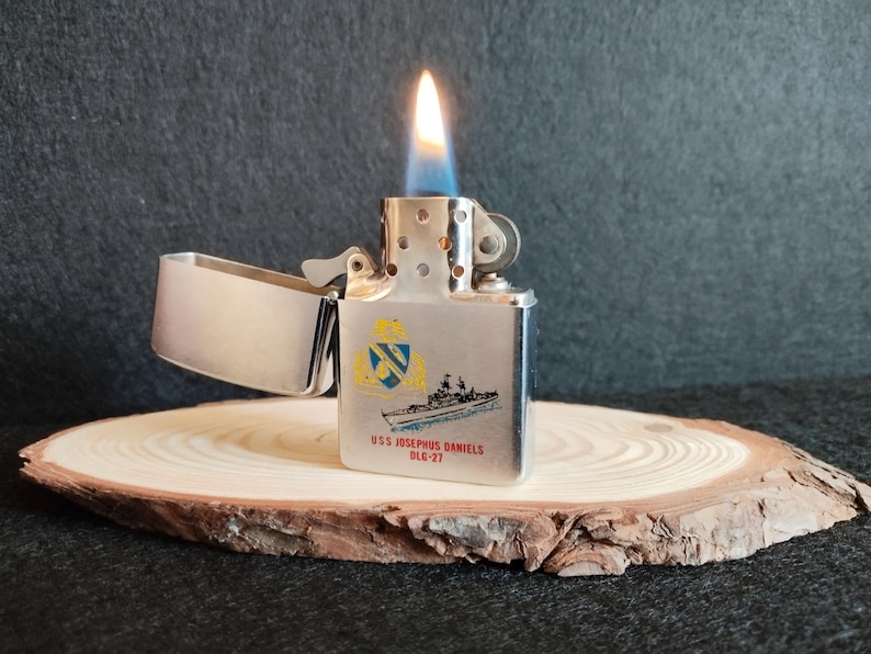 Puede incluir: Un encendedor de metal plateado con una llama, sobre una rodaja de madera. El encendedor presenta un dise&ntilde;o de barco y el texto "USS JOSEPHUS DANIELS DLG-27" en negro y azul.