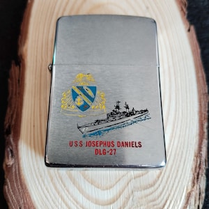 Puede incluir: Un encendedor plateado con una ilustraci&oacute;n detallada de un barco y un escudo. El texto "USS JOSEPHUS DANIELS DLG-27" est&aacute; impreso debajo de la imagen del barco. El encendedor se muestra sobre una superficie de madera.
