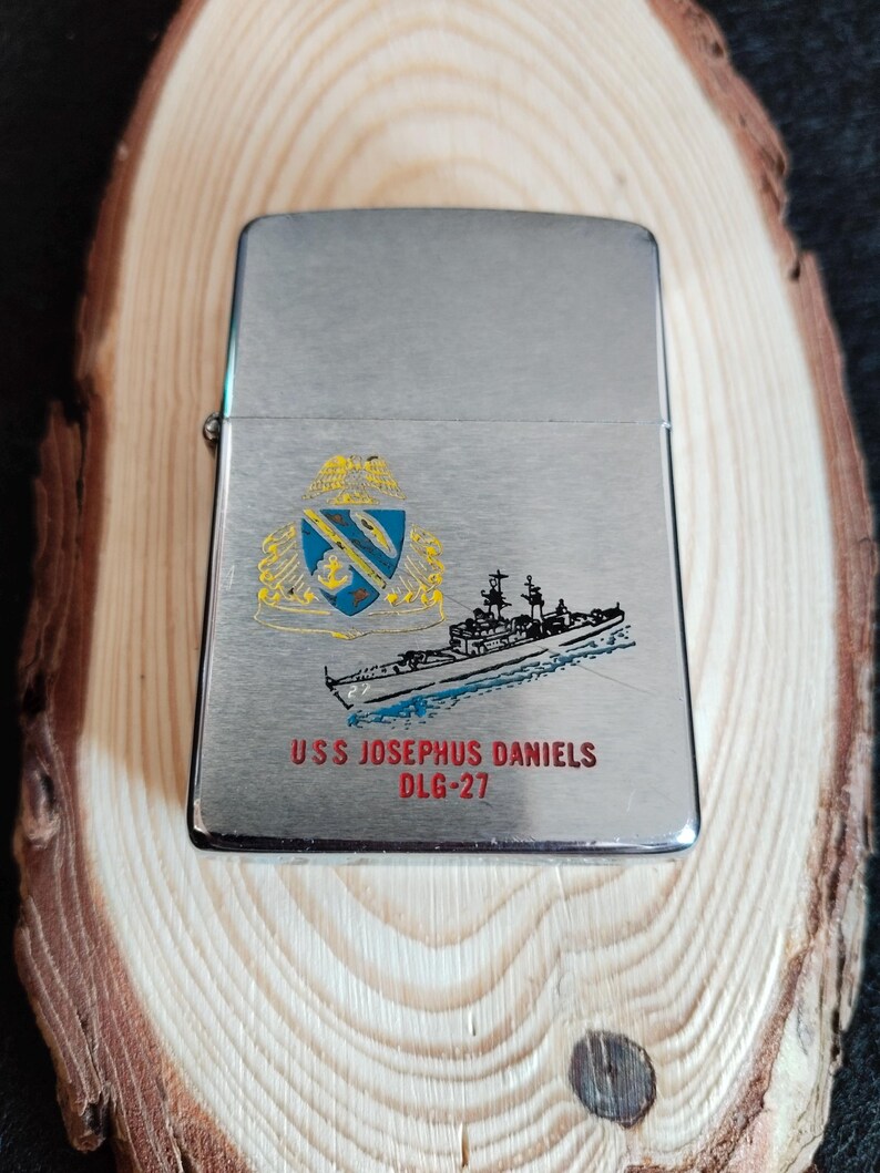 Puede incluir: Un encendedor plateado con una ilustraci&oacute;n detallada de un barco y un escudo. El texto "USS JOSEPHUS DANIELS DLG-27" est&aacute; impreso debajo de la imagen del barco. El encendedor se muestra sobre una superficie de madera.