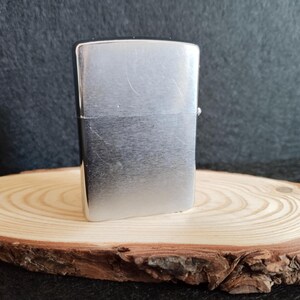 Puede incluir: Un encendedor de metal plateado, de forma rectangular con esquinas redondeadas y una tapa con bisagras. Se encuentra sobre una rodaja de madera redonda, con un fondo oscuro.