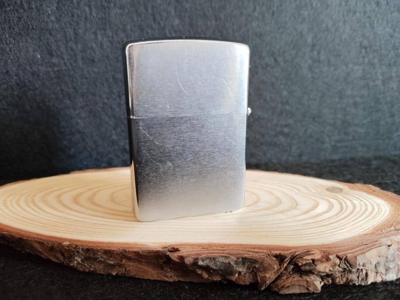 Puede incluir: Un encendedor de metal plateado, de forma rectangular con esquinas redondeadas y una tapa con bisagras. Se encuentra sobre una rodaja de madera redonda, con un fondo oscuro.