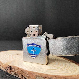 Może przedstawiać: Srebrna zapalniczka z otwartą pokrywą, spoczywająca na drewnianej podstawie. Zapalniczka posiada niebiesko-biały emblemat z napisem "STANAVFORLANT". Tło jest ciemne.