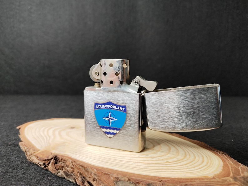 Może przedstawiać: Srebrna zapalniczka z otwartą pokrywą, spoczywająca na drewnianej podstawie. Zapalniczka posiada niebiesko-biały emblemat z napisem "STANAVFORLANT". Tło jest ciemne.