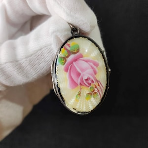 Può includere: Un medaglione di forma ovale con una rosa rosa dipinta a mano e foglie verdi su uno sfondo color crema caratterizzato da una fine texture guilloché radiante. Il medaglione è incorniciato da un bordo in metallo scuro e ha un gancio argentato.