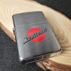 Peut inclure: Un briquet argent&eacute; avec le mot "Manitowoc" imprim&eacute; en noir sur un cercle rouge. Le briquet rectangulaire a des bords arrondis et une finition brillante. Le briquet ferm&eacute; repose sur une surface en bois.