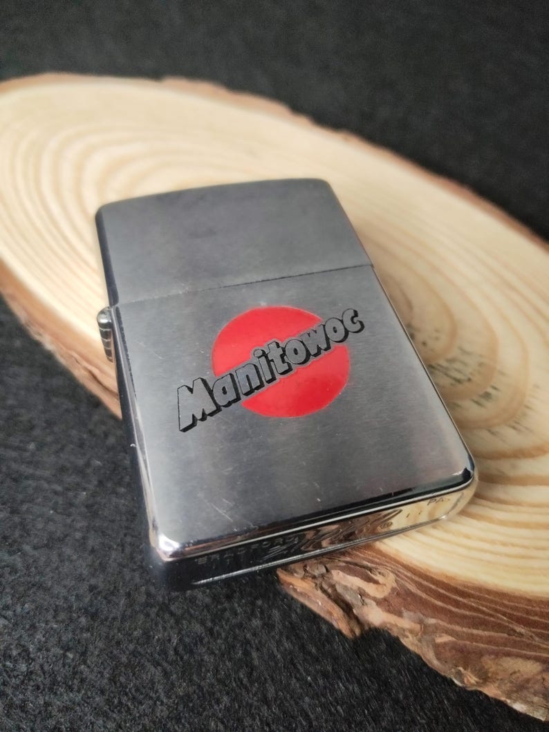 Peut inclure: Un briquet argent&eacute; avec le mot "Manitowoc" imprim&eacute; en noir sur un cercle rouge. Le briquet rectangulaire a des bords arrondis et une finition brillante. Le briquet ferm&eacute; repose sur une surface en bois.