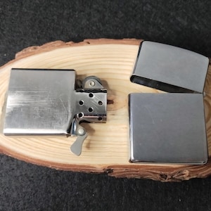 Peut inclure: Un briquet en m&eacute;tal argent&eacute; avec un couvercle &agrave; charni&egrave;re ouvert, r&eacute;v&eacute;lant la molette &agrave; silex et la m&egrave;che. Deux &eacute;tuis de briquet en m&eacute;tal argent&eacute; suppl&eacute;mentaires sont &eacute;galement pr&eacute;sents. Les articles sont pr&eacute;sent&eacute;s sur une tranche de bois naturelle.