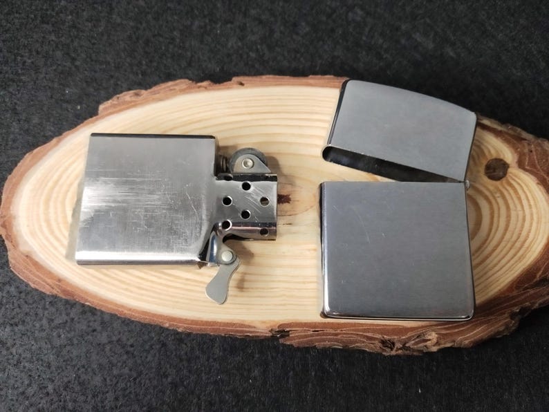 Peut inclure: Un briquet en m&eacute;tal argent&eacute; avec un couvercle &agrave; charni&egrave;re ouvert, r&eacute;v&eacute;lant la molette &agrave; silex et la m&egrave;che. Deux &eacute;tuis de briquet en m&eacute;tal argent&eacute; suppl&eacute;mentaires sont &eacute;galement pr&eacute;sents. Les articles sont pr&eacute;sent&eacute;s sur une tranche de bois naturelle.