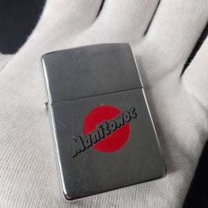 Peut inclure: Un briquet argent&eacute; avec le mot "Manitowoc" en &eacute;criture noire sur un cercle rouge. Le briquet rectangulaire a des bords arrondis et un couvercle &agrave; charni&egrave;re. Le briquet est tenu dans un gant blanc.