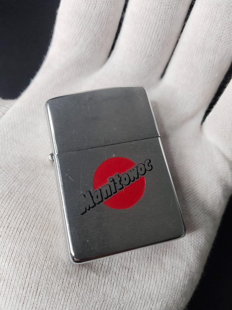 Peut inclure: Un briquet argent&eacute; avec le mot "Manitowoc" en &eacute;criture noire sur un cercle rouge. Le briquet rectangulaire a des bords arrondis et un couvercle &agrave; charni&egrave;re. Le briquet est tenu dans un gant blanc.