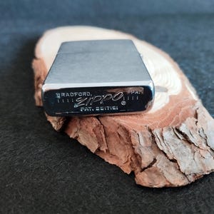 Puede incluir: Un encendedor Zippo plateado con el texto "BRADFORD, PA." y "ZIPPO" impreso en la parte inferior. El encendedor est&aacute; sobre un trozo de madera con una superficie rugosa y texturizada. El fondo es de color oscuro y liso.