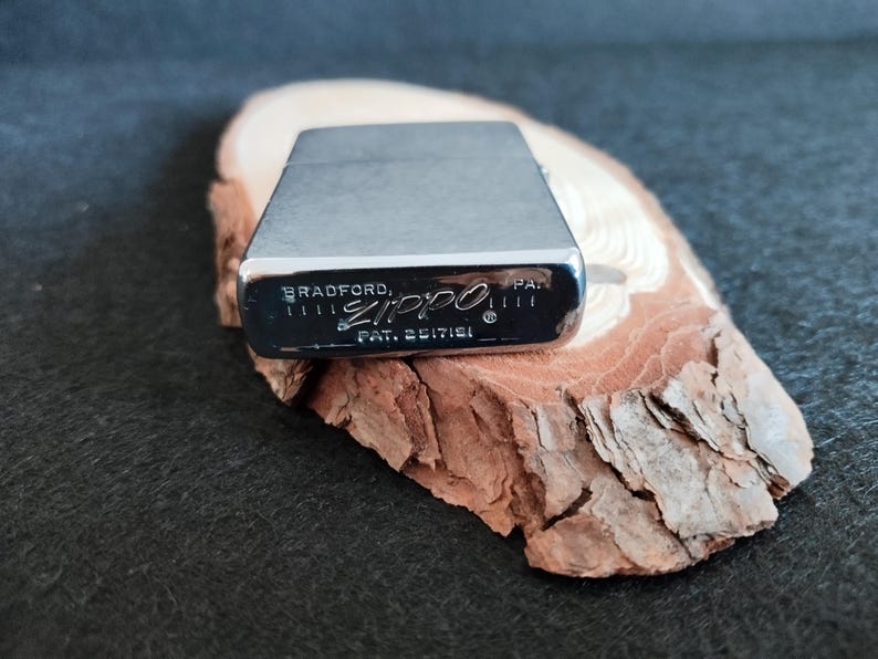 Puede incluir: Un encendedor Zippo plateado con el texto "BRADFORD, PA." y "ZIPPO" impreso en la parte inferior. El encendedor est&aacute; sobre un trozo de madera con una superficie rugosa y texturizada. El fondo es de color oscuro y liso.