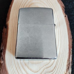 Puede incluir: Un encendedor rectangular plateado con un acabado de metal cepillado. El encendedor est&aacute; sobre una secci&oacute;n transversal de un tronco de madera de color claro, mostrando los patrones naturales de la veta. El fondo es oscuro.