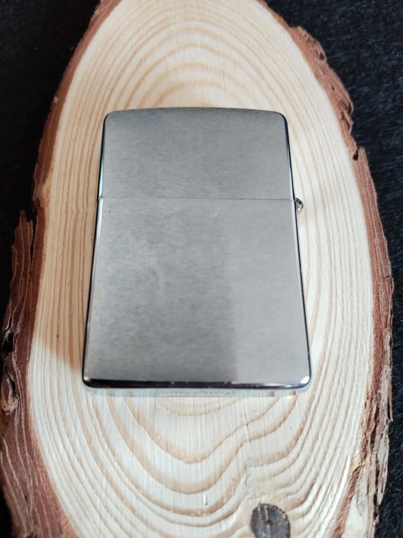 Puede incluir: Un encendedor rectangular plateado con un acabado de metal cepillado. El encendedor est&aacute; sobre una secci&oacute;n transversal de un tronco de madera de color claro, mostrando los patrones naturales de la veta. El fondo es oscuro.