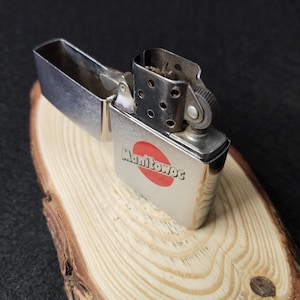 Peut inclure: Un briquet argent&eacute; ouvert, avec le mot "Manitowoc" et un cercle rouge imprim&eacute;s. Il est pos&eacute; sur une surface en bois clair, sur fond sombre.